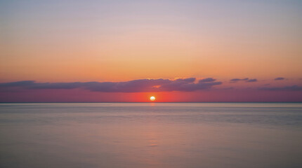 Fototapeta premium A serene sunset paints the sky with hues of orange and pink, casting a warm glow over the tranquil ocean. The sun dips below the horizon, reflected on the water's surface.