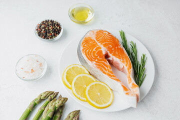 Raw salmon steak with lemon slices, rosemary, asparagus and spices on white plate over light...