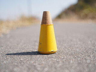 A bright yellow traffic cone with a wooden top stands on an asphalt road, casting a shadow on a sunny day