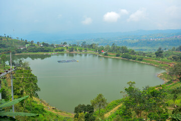 Fototapeta premium A tranquil lake nestled within a verdant valley, surrounded by lush green hills and distant mountains. Related to rural landscapes, nature conservation, travel to quiet destinations, scenic beauty