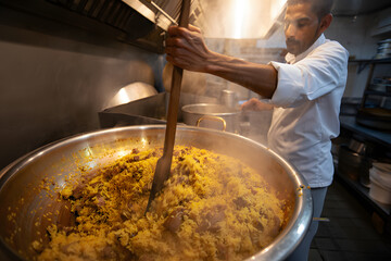 Chef stirring aromatic saffron rice and meat in large commercial kitchen pot.