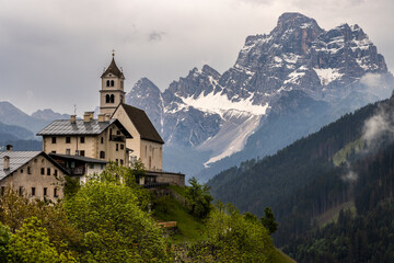 Fototapeta premium Church of Colle Santa Lucia village perched on a cliff top with a dramatic misty and moody backdrop of Monte Pelmo in the Dolomites, Italy