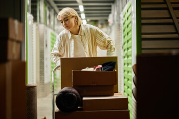 Caucasian woman pushing cart loaded with cardboard boxes in storage facility, appearing focused...