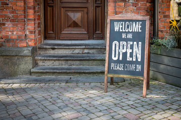 WELCOME WE ARE OPEN PLEASE COME IN. Entrance Signboard on Cobblestone Street