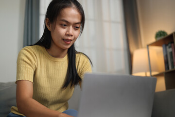 Portrait of Young Asian woman sitting on sofa working on laptop in cozy living room at home. Remote...