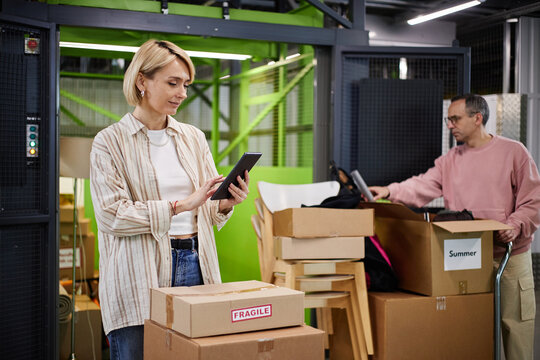 Caucasian middle aged woman using digital tablet while standing among cardboard boxes in storage facility, man organizing items in background
