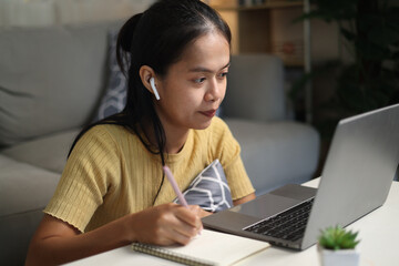 Portrait of Young Asian woman sits on the floor taking online tutoring lessons on a laptop in...
