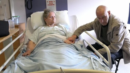 Tender moment as an elderly husband sits beside his ill wife in a hospital bed, gently holding her hand in a supportive and caring gesture during a medical recovery or palliative care setting - Powered by Adobe