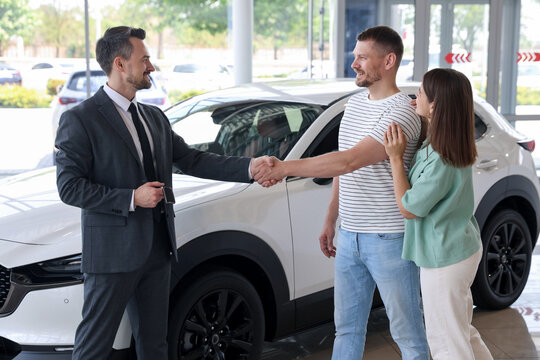 Salesman shaking hands with happy family near new car in salon