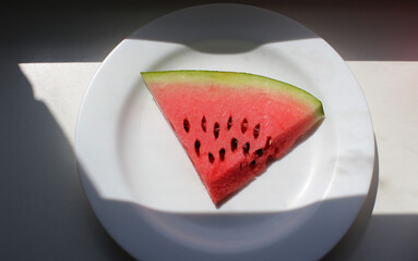 A slice of watermelon on a plate on a windowsill with shade from the sun 
