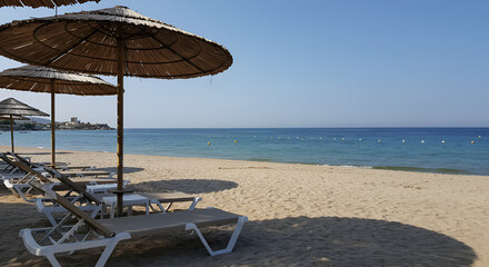 Beach scene with straw umbrellas casting shadows on empty sunbeds, overlooking a calm blue sea under a clear sky.