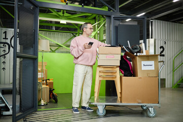 Middle aged Caucasian man standing beside storage unit holding smartphone, organizing cardboard...