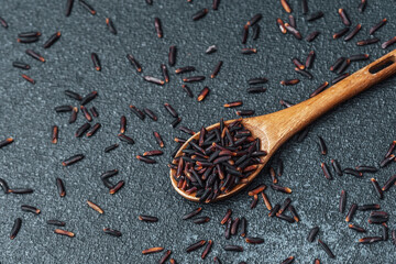 Wooden spoon with black rice on dark background with scattered grains