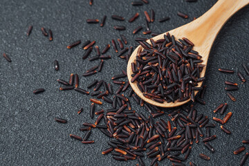 Wooden spoon filled with black rice and spilled grains on dark background