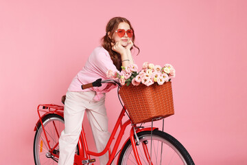 Beautiful teenage girl in sunglasses posing on bicycle with basket of flowers against pink...