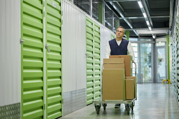 Middle aged Caucasian man pushing cart loaded with cardboard boxes through corridor of self storage facility, looking ahead while transporting items, storage units visible on both sides
