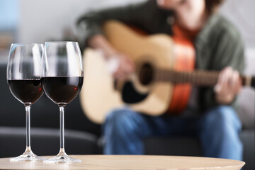 Glasses with red wine on table and man playing guitar indoors, selective focus