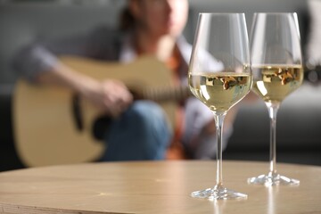 Glasses with white wine on table and man playing guitar indoors, selective focus