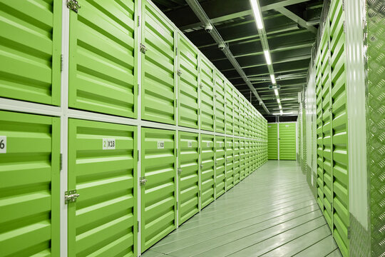 Row of green storage unit doors lining corridor in modern self storage facility, metal walls and ceiling visible, fluorescent lights illuminating clean organized environment