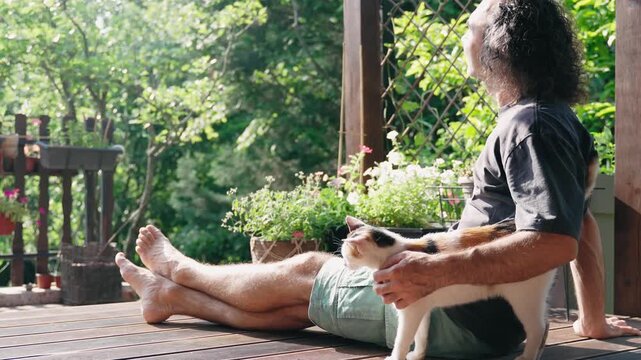 A gentle man strokes a cat while sitting on the sunny terrace of a country house
