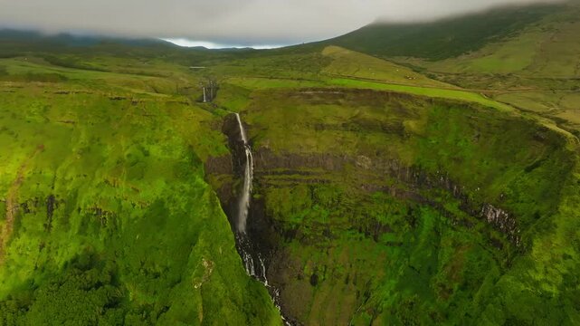 Aerial view of the Cascata do Poco do Bacalhau waterfall plunging down lush green cliffs, creating a mesmerizing contrast with the dark rock, Faj&atilde; Grande, Azores, Portugal.