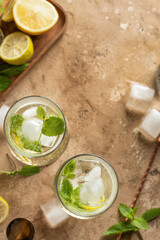 Mojito long rum drink with fresh mint, lemon juice, ice cubes and soda on brown textured background. Summer party cocktails with copy space, top view