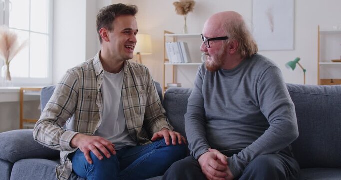 Elderly father and adult son sitting together on sofa in living room and having joyful conversation. Happy family smiling while discussing funny issues and sharing stories during dialogue at home.