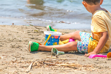 Toddler playing with sand toys on the beach. Child on summer vacation. Tourist boy at the seaside. Faceless kid playing with bucket and shovel set. Outdoor activity. Alone. Horizontal photo.