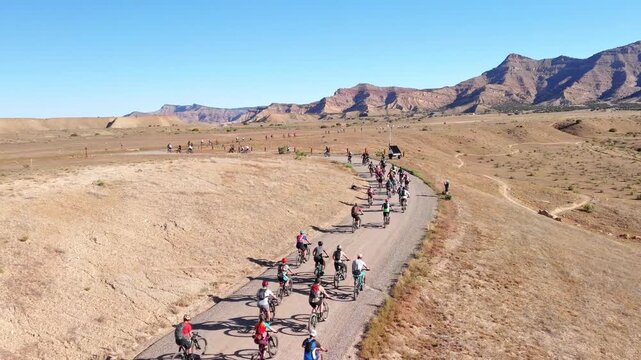 Fruita, United States - 28 July 2025: Aerial view of a group of cyclists riding down a winding road through arid terrain, with majestic mountains in the distance.