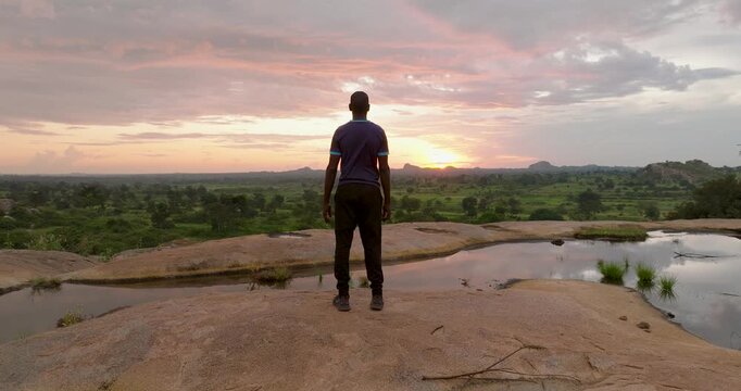 Nyero, Uganda - 06 December 2025: Aerial view of a man standing on a rocky outcrop overlooking a lush landscape under a serene sunset sky.