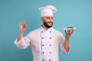 Happy confectioner in uniform holding tart of blueberries and showing ok gesture on light blue background