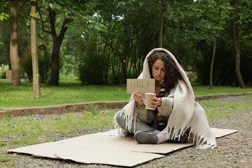 Homeless woman with sign Help and paper cup on carton in park, space for text