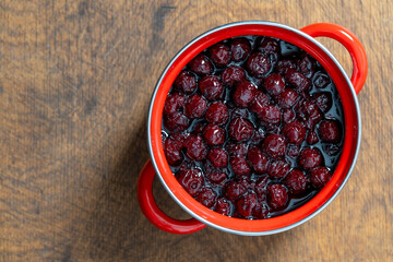 Delicious cherry jam in a red saucepan on a wooden table, closeup, top view. Summer preparation and preservation of berries for the winter, home cooking