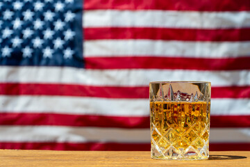 Glass of whiskey on wooden table against the backdrop of an American flag, closeup. Bourbon, an American whiskey, is often associated with the United States and is considered the national drink