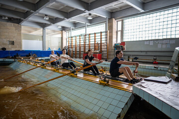 Exhausted athletes rapidly rowing on simulator, working on synchronized techniques. Rowers hone...