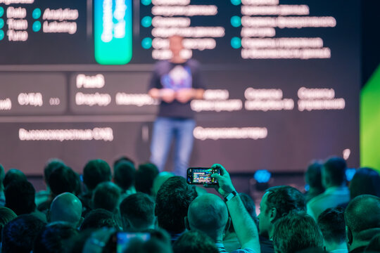 Crowd attending business event while recording presenter on stage with phone in hand.