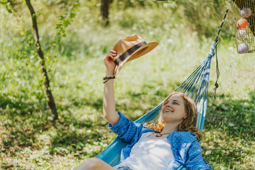 Happy young blonde woman with long curly hair in hat swinging on hammock in summer garden