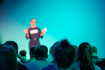 Person speaking in front of audience against turquoise background. Seminar or conference setting.