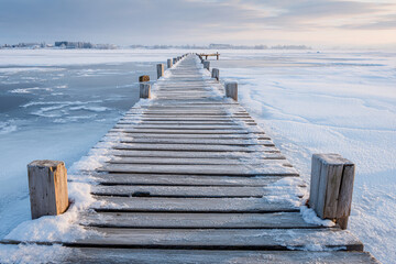 Obraz premium Wooden pier covered in snow leading to frozen lake