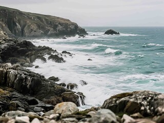 Waves from a moody, stormy sea crash into dark, jagged rocks along a remote coastline. The overcast sky and raw ocean power create a dramatic and untamed scene 