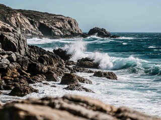 Fototapeta premium Strong waves crash against rugged rocks along a dramatic coastal shoreline under a clear blue sky. The turbulent sea and textured cliffs evoke a sense of raw power and untamed nature