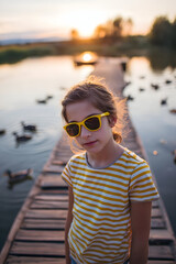 Young girl wearing yellow sunglasses stands on wooden dock at sunset with ducks swimming in the water, enjoying a peaceful evening outdoors