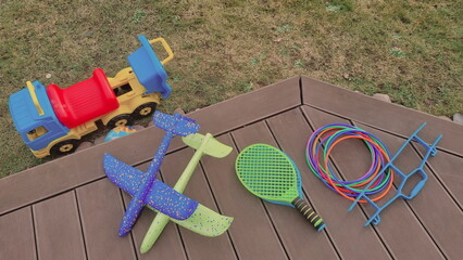 Backyard photo with a pile of outdoor toys for kids. A bunch of sports toys are lying on a wooden patio. Tennis rackets, plastic hoops, planes made of foam, and toy vehicle are beloved toys for boys. 