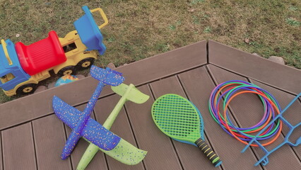 Backyard photo with a pile of outdoor toys for kids. A bunch of sports toys are lying on a wooden patio. Tennis rackets, plastic hoops, planes made of foam, and toy vehicle are beloved toys for boys. 