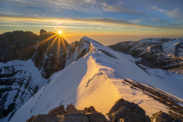 Aerial view of the sun casting golden light on snow-capped mountain peaks, creating a breathtaking panorama, as seen from above, Location Unknown.