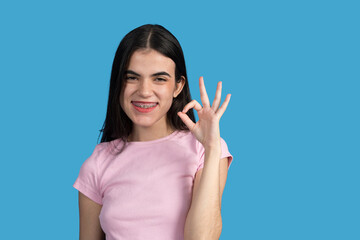 Young woman with braces smiling and gesturing ok sign on blue background