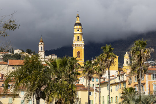 View of the vibrant yellow bell tower piercing through the clouds, framed by lush palms and terracotta rooftops, Menton, Provence-Alpes-C&Atilde;&acute;te d'Azur, France.