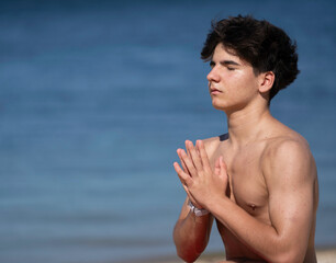 Close up side view of shirtless young man practicing yoga and meditating on the beach, enjoying the...