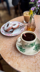 donuts dusted with powdered sugar on a floral plate, served with iced coffee and hot black coffee on a vintage-style café table.