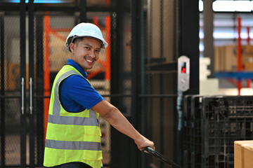 An Asian Male warehouse worker in a safety vest and helmet operating a pallet jack, demonstrating...